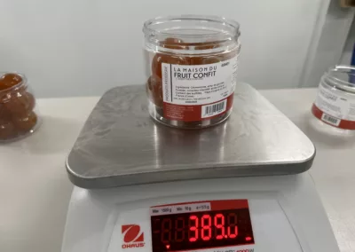 conditionnement Aptunion : Un pot de fruits confits pesé sur une balance électronique, affichant un poids de 389 grammes. L’étiquette indique "La Maison du Fruit Confit